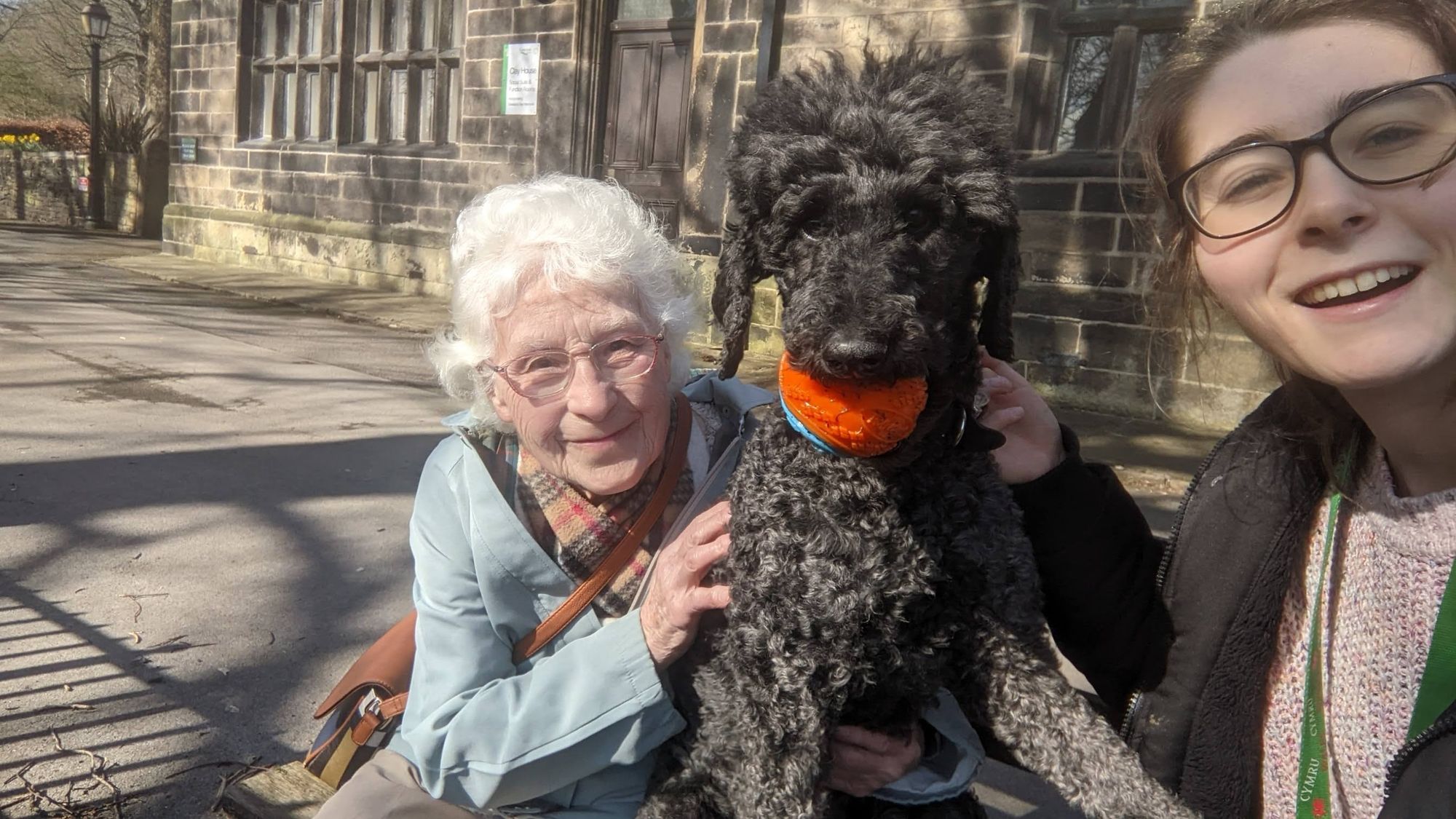 A photo showing Iris and Molly surrounding a poodle named Dora, who is holding an orange ball in her mouth